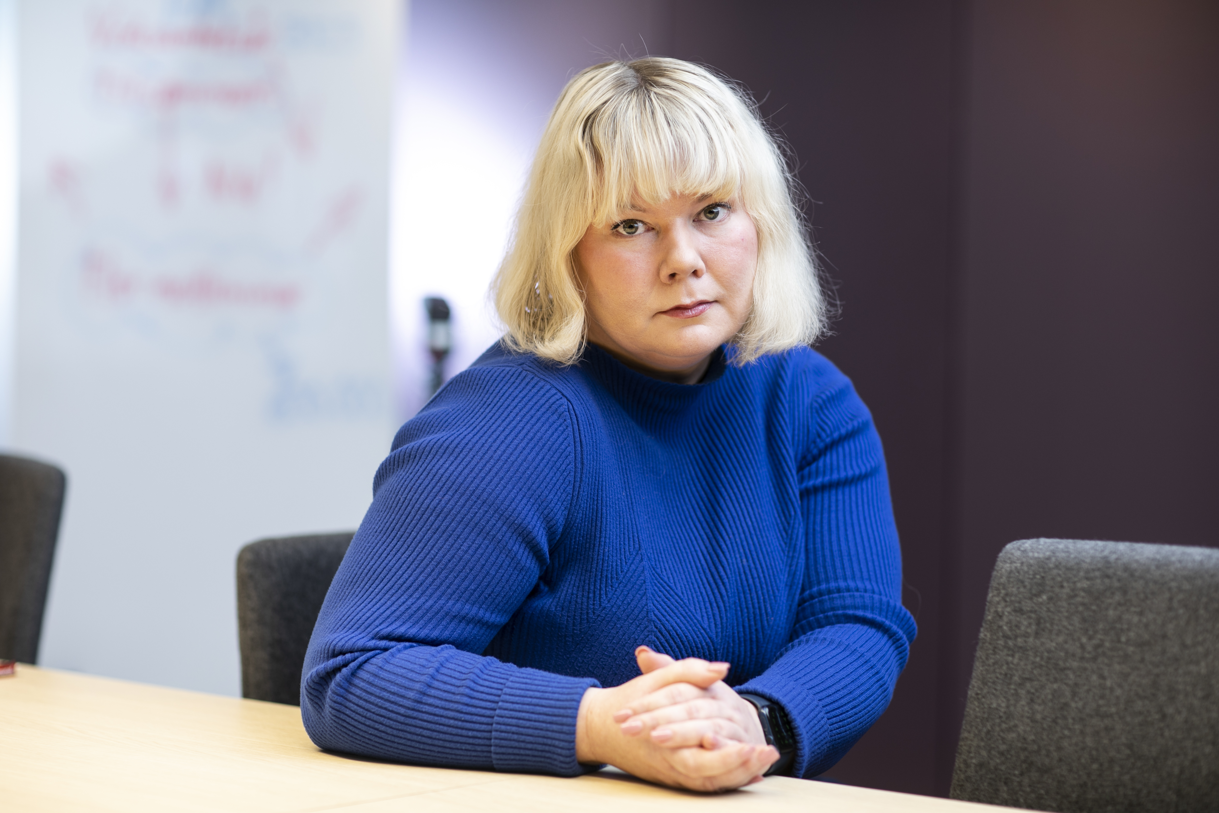 Mari Huupponen med blont hår, iklädd en blå tröja, sitter vid ett bord och har händerna knäppta. Hon tittar direkt i kameran.