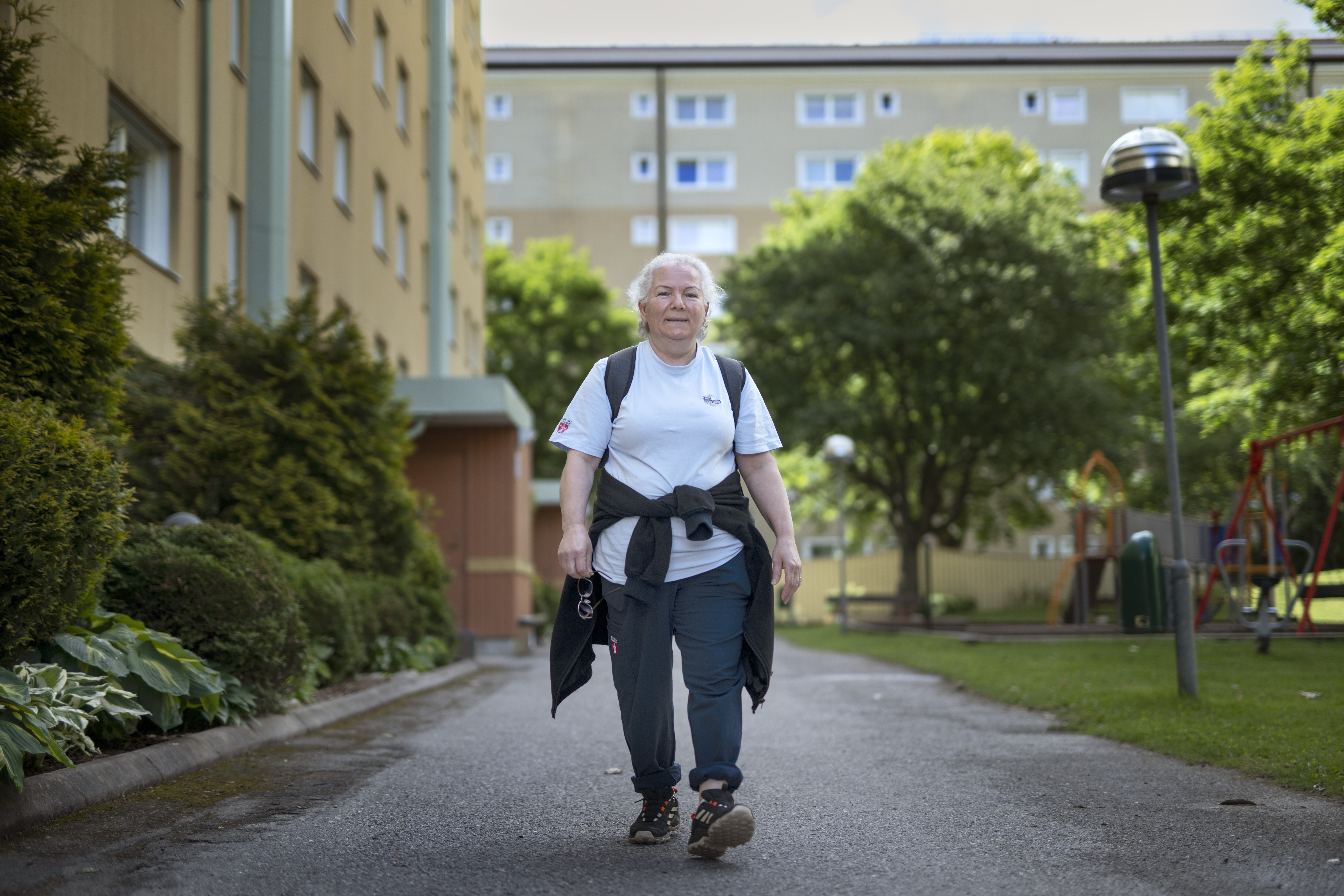 Marin Gabriel i ledig klädsel går längs en gångväg i ett bostadsområde med växter, en byggnad och lekutrustning i bakgrunden.