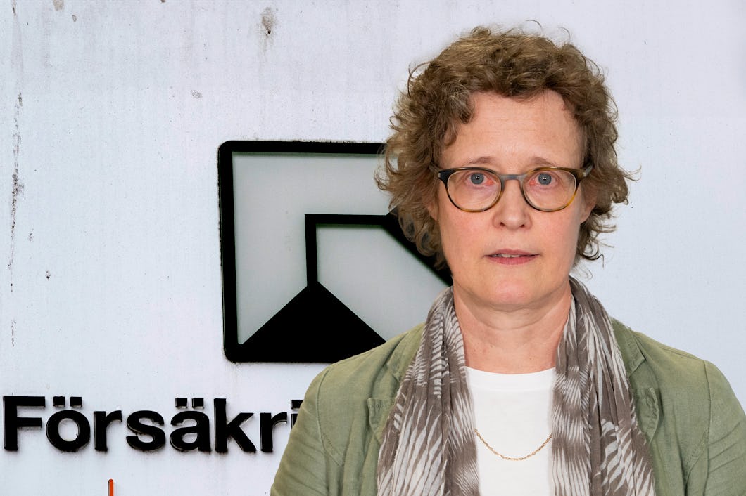 En person med kort lockigt hår och glasögon, iklädd en scarf, står framför en skylt med texten "Försäkringskassan".