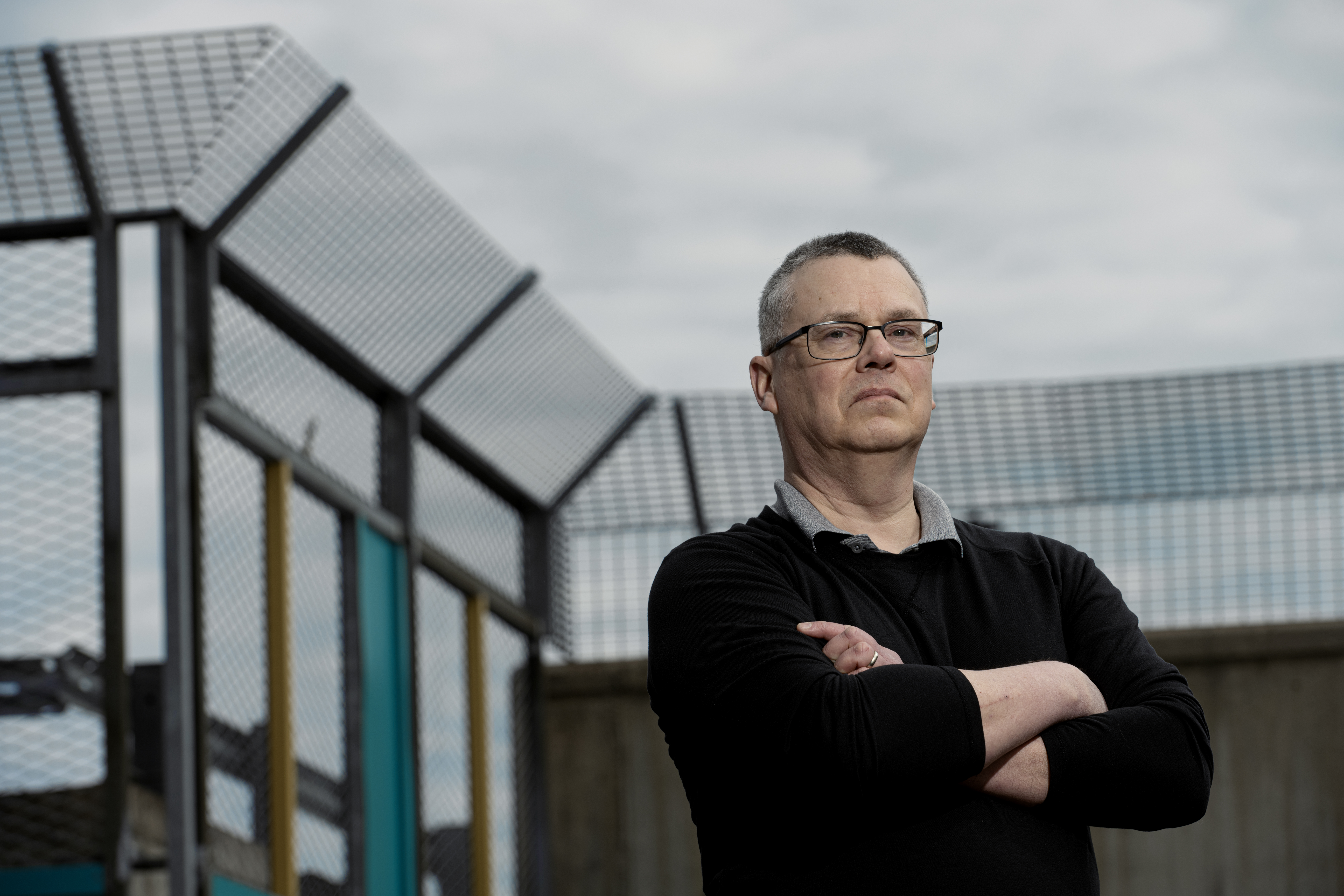 https://images.lomediehuPatrik Nylund med glasögon och korslagda armar står utomhus nära en inhägnad struktur, under en molnig himmel.