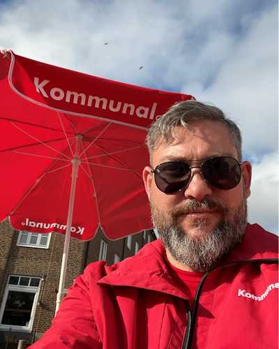Philip Corter med grått hår, solglasögon och skägg står framför ett rött paraply märkt "Kommunal". Han har på sig en röd jacka. Himlen är delvis molnig och några fåglar syns i bakgrunden.