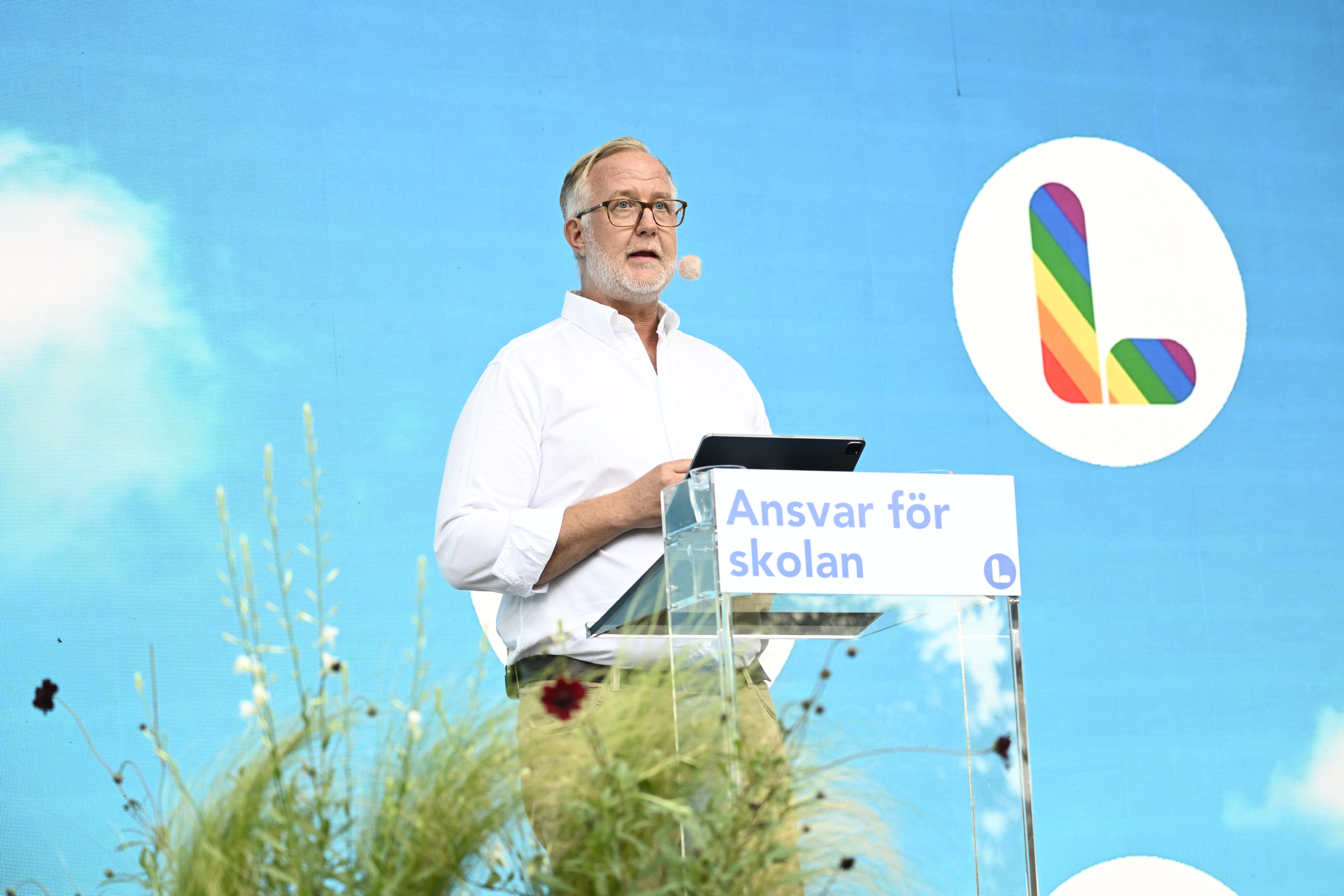 Johan Pehrson talar vid ett podium med en skylt som lyder "Ansvar för skolan" framför en blå bakgrund med en regnbågsfärgad L-logotyp.