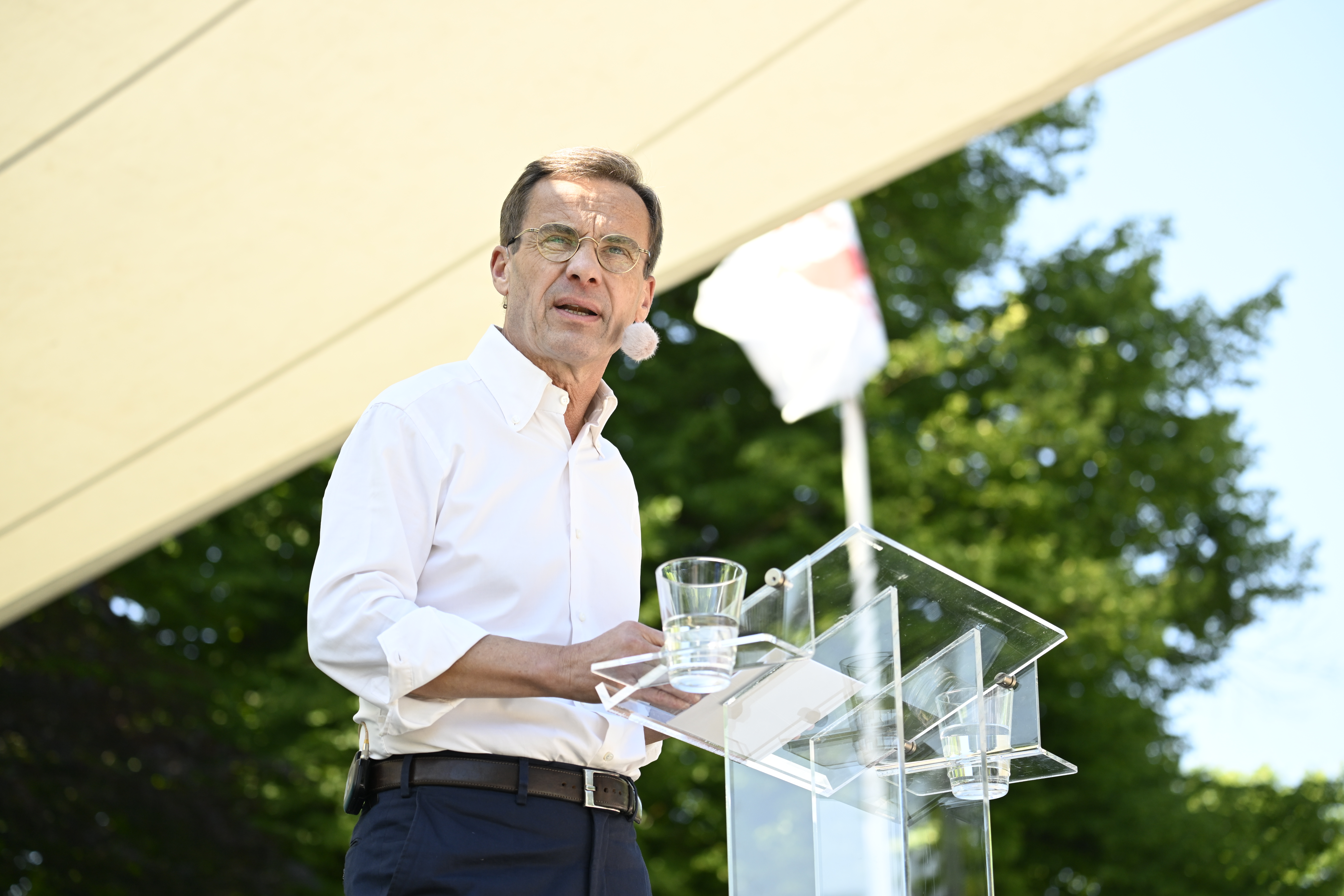 En man med glasögon och vit skjorta står bakom ett genomskinligt podie och talar i ett headset-mikrofon. Ett glas vatten står på podiet. I bakgrunden syns en flagga och växter.