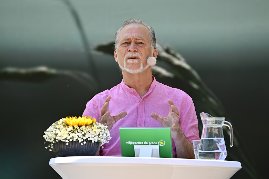En man står bakom ett podium med ett headset-mikrofon och en rosa skjorta, gestikulerande med händerna. På podiet finns en vas med blommor bredvid en vattenkanna.