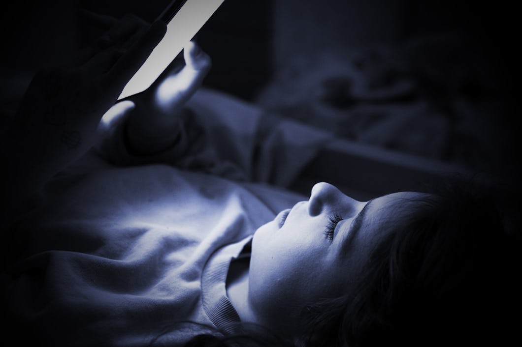 En person ligger i sängen i mörkret, belyst av ljuset från telefon som håller i handen.