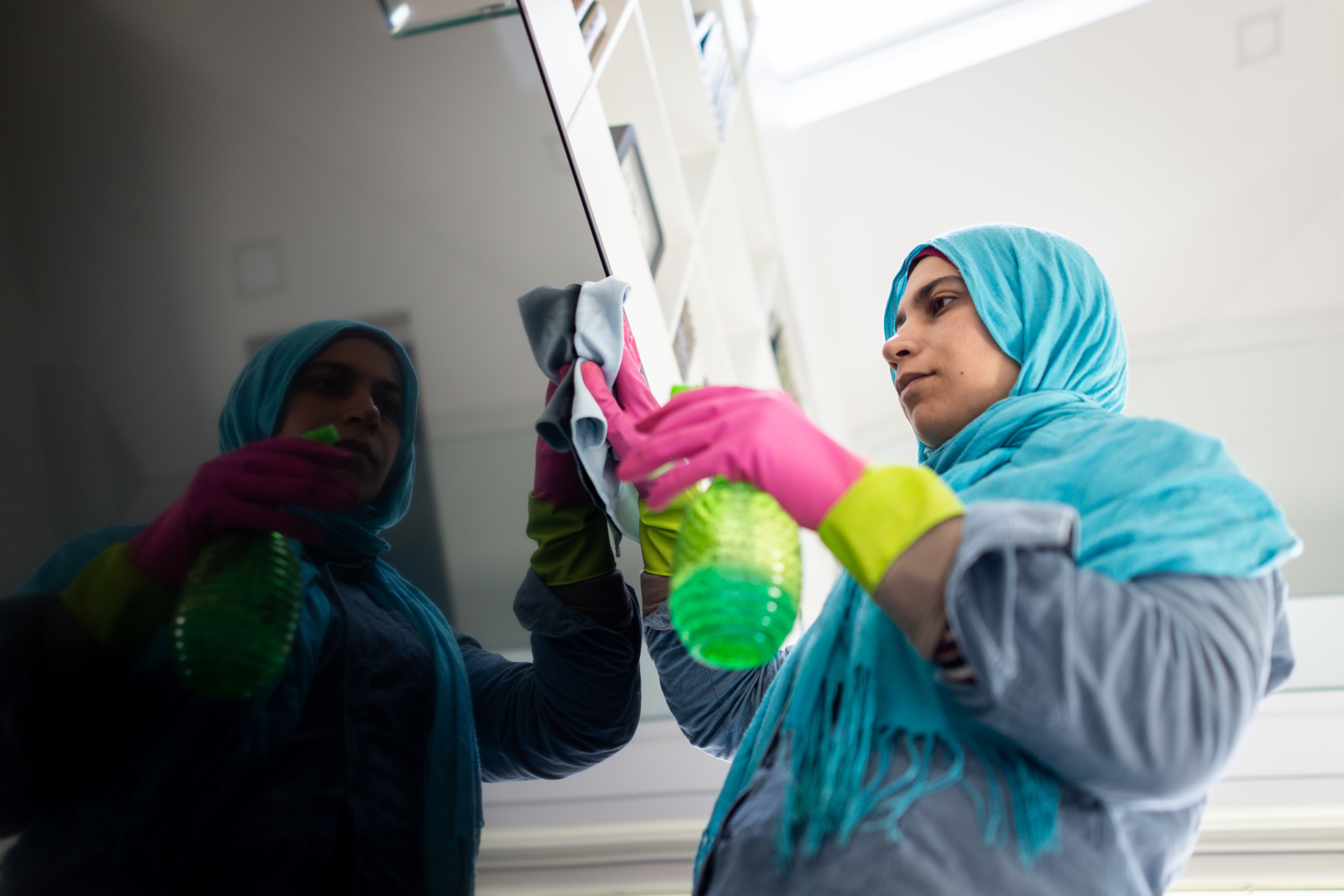 En person med en blå huvudduk och rosa handskar rengör en spegelyta med en trasa och en grön sprayflaska.