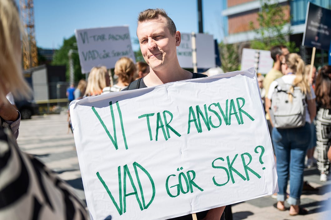 En person håller ett plakat med texten "Vi tar ansvar, vad gör SKR?" på en protest. Andra människor och skyltar syns i bakgrunden. Stadsmiljö med moderna byggnader och klar himmel.