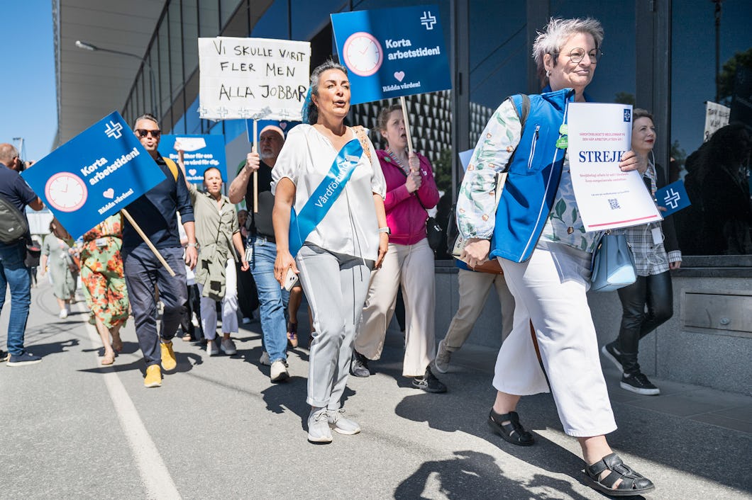 En grupp människor deltar i en protest utomhus och håller upp skyltar och banderoller som kräver kortare arbetstid och bättre villkor. Några bär västar som visar deras stöd för saken.