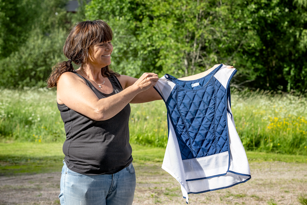 Tina Persson står utomhus och håller upp ett blått och vitt vadderat plagg, en kylväst. Hon har på sig en svart linne och blå jeans. I bakgrunden syns gröna träd och gräs.