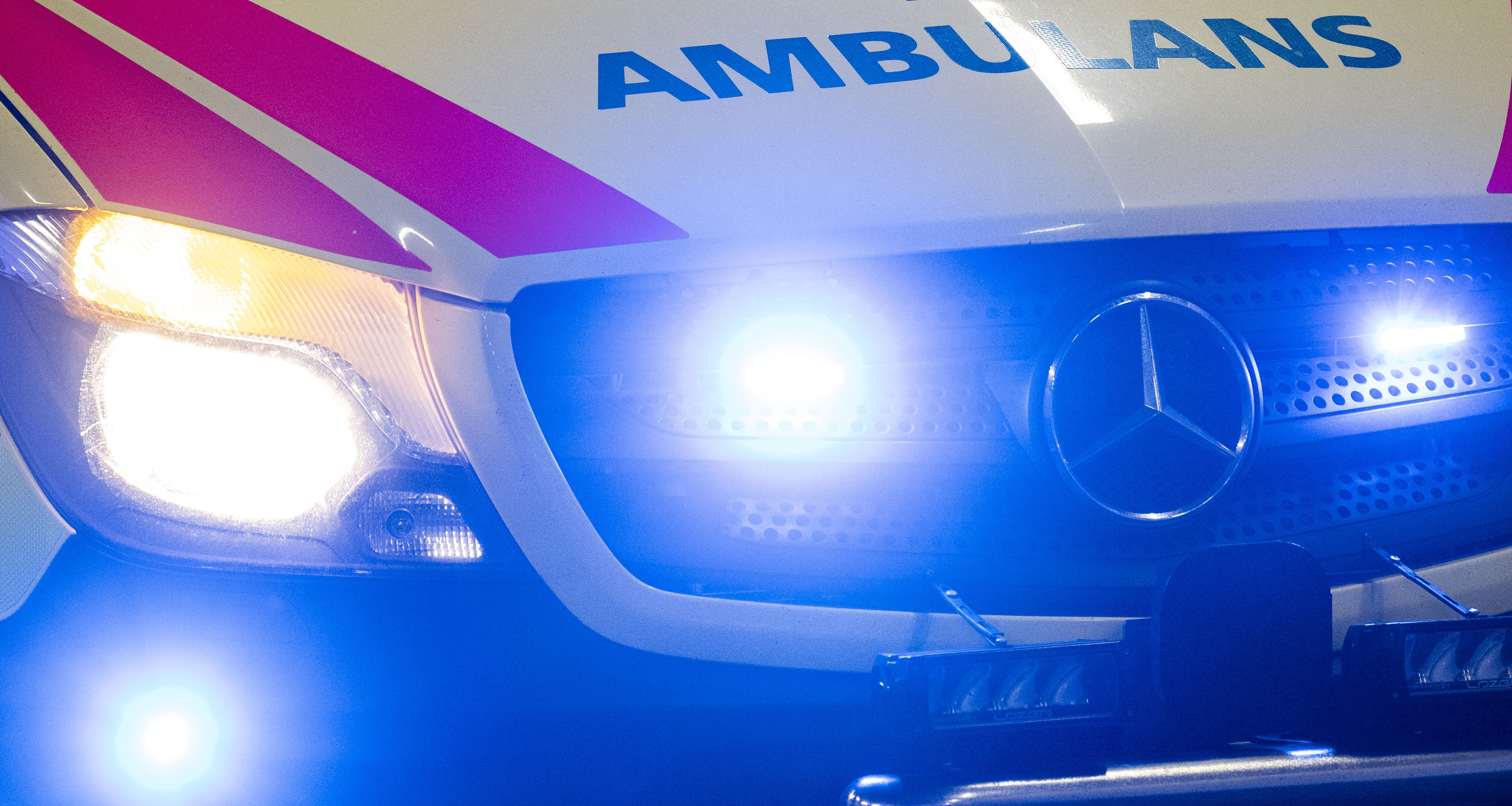 Närbild på en Mercedes-Benz ambulans med tända strålkastare och blåa varningsljus aktiverade.