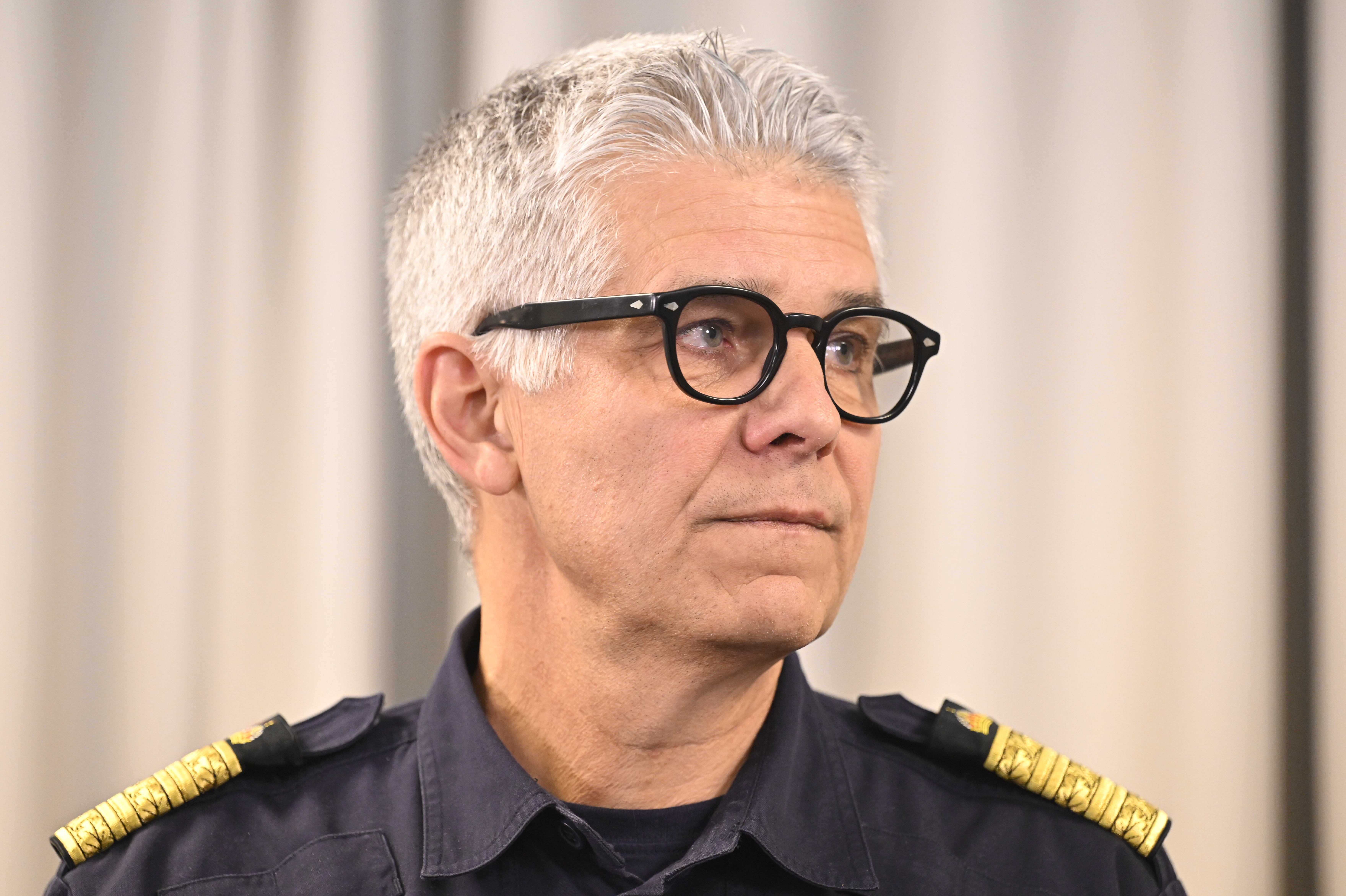 En person med kort grått hår och glasögon, iförd en mörk uniform med epåletter, står framför ett ljusfärgat draperi.