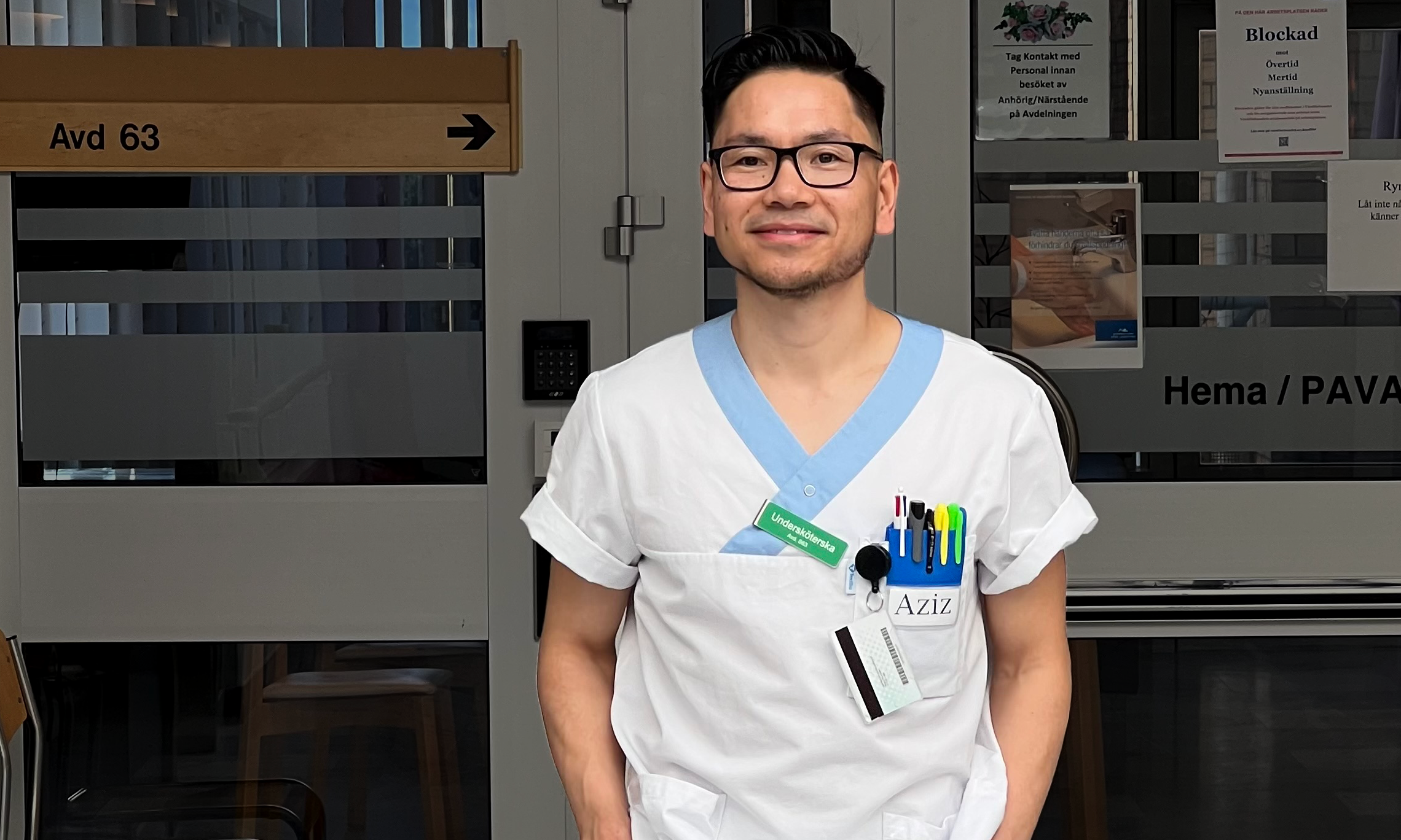 Aziz Ehsani klädd i vit uniform står framför en dörr med skyltarna "Hema/PAVA" och "Avd 63." Uniformen har en namnskylt med texten "Aziz," och flera pennor är fastklämda på bröstfickan.