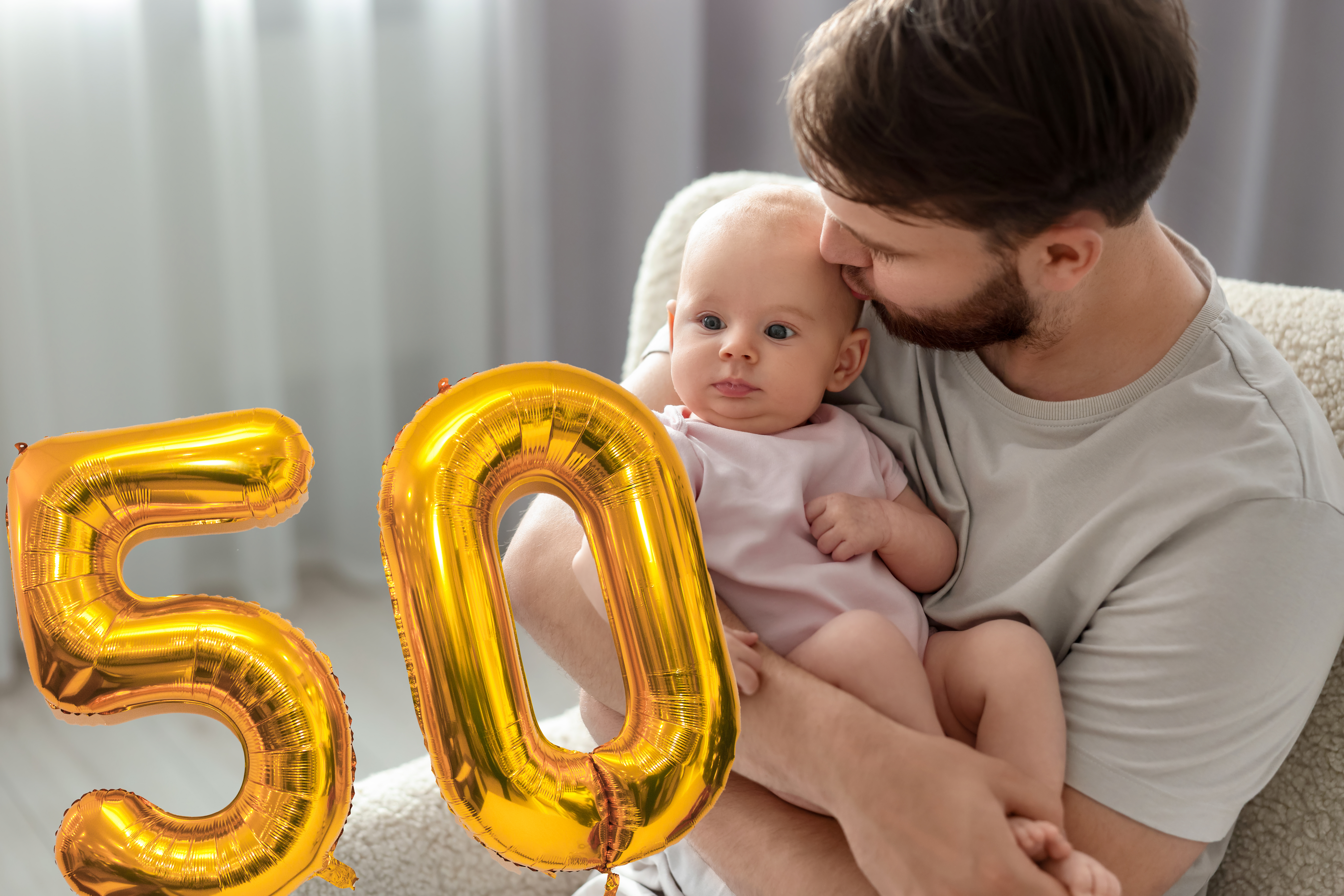 En man sitter på en stol och håller en baby. I förgrunden finns två guldfolieballonger som formar siffran "50".