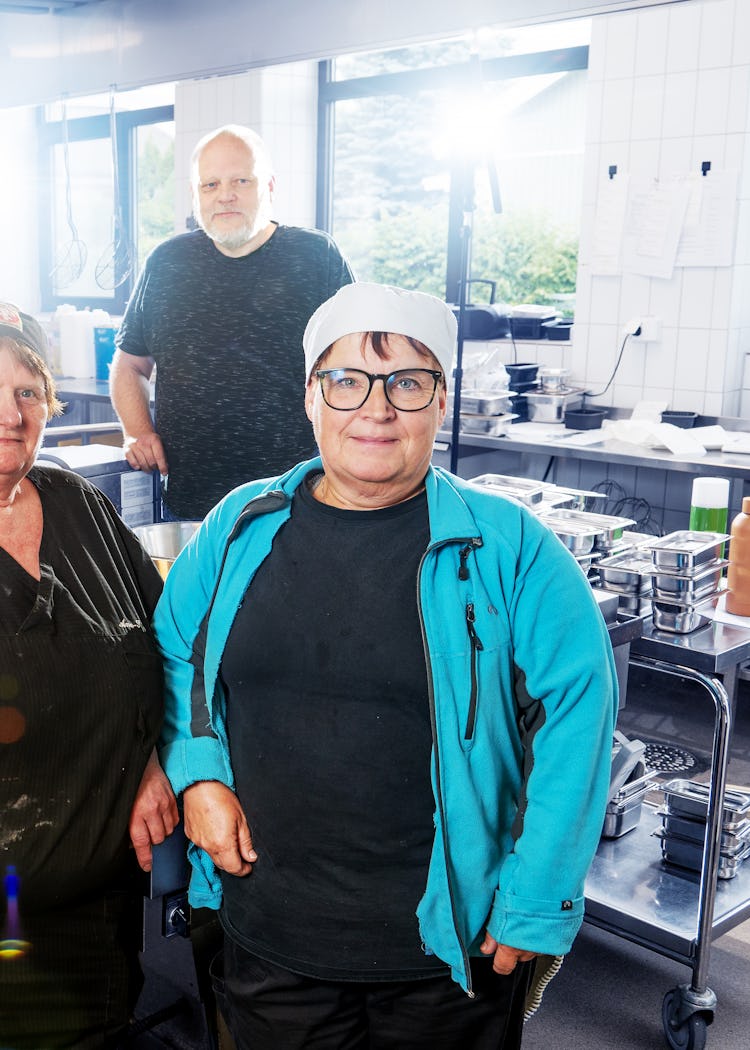 Fyra personer står i ett kommersiellt kök och poserar för ett foto. Tre kvinnor och en man bär lediga kläder och arbetskläder, omgivna av olika köksutrustningar och apparater.