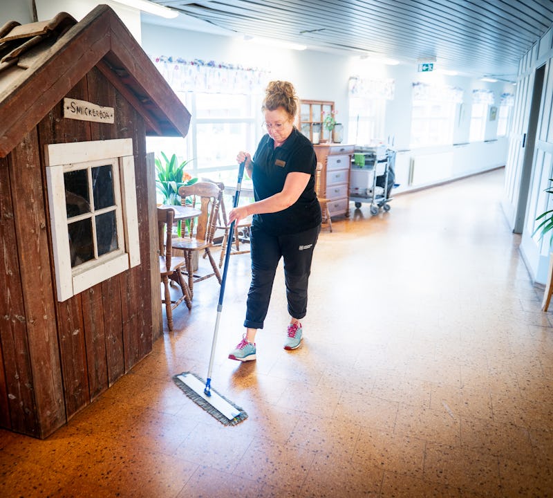 En person moppar golvet i en korridor bredvid ett litet trähus för lek samt några bord och stolar.