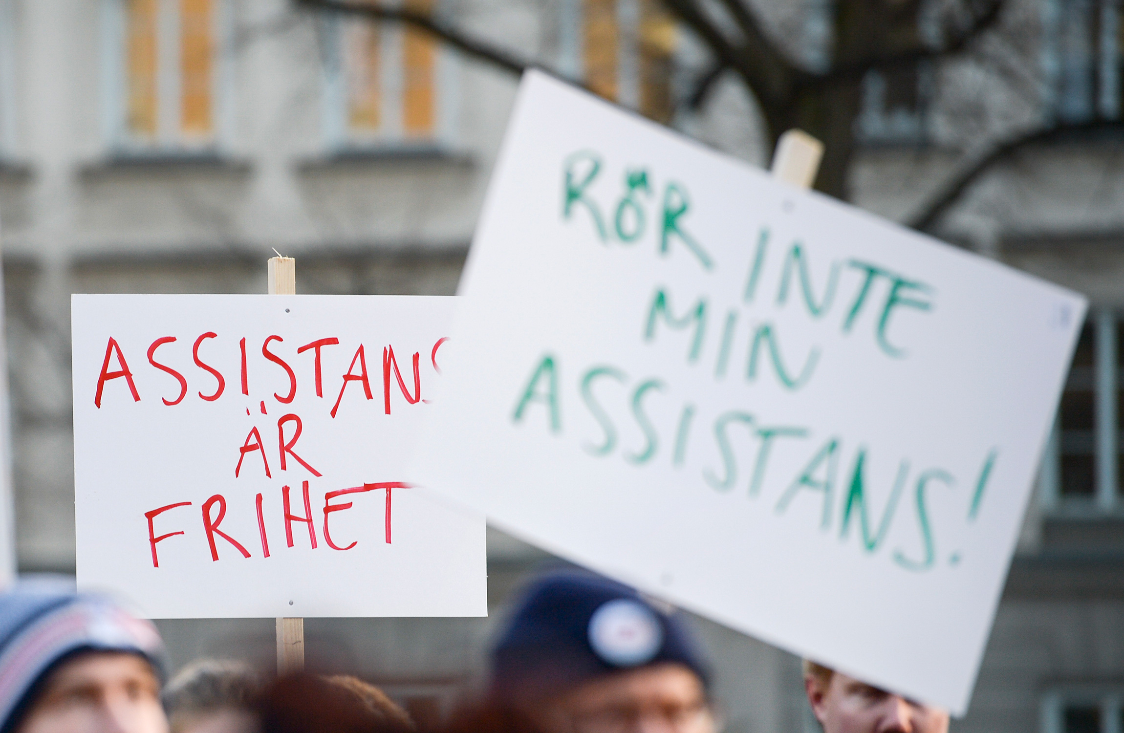 Två demonstrationsskyltar på svenska hålls upp av deltagare vid en demonstration. En skylt lyder "ASSISTANS ÄR FRIHET" och den andra lyder "RÖR INTE MIN ASSISTANS!".