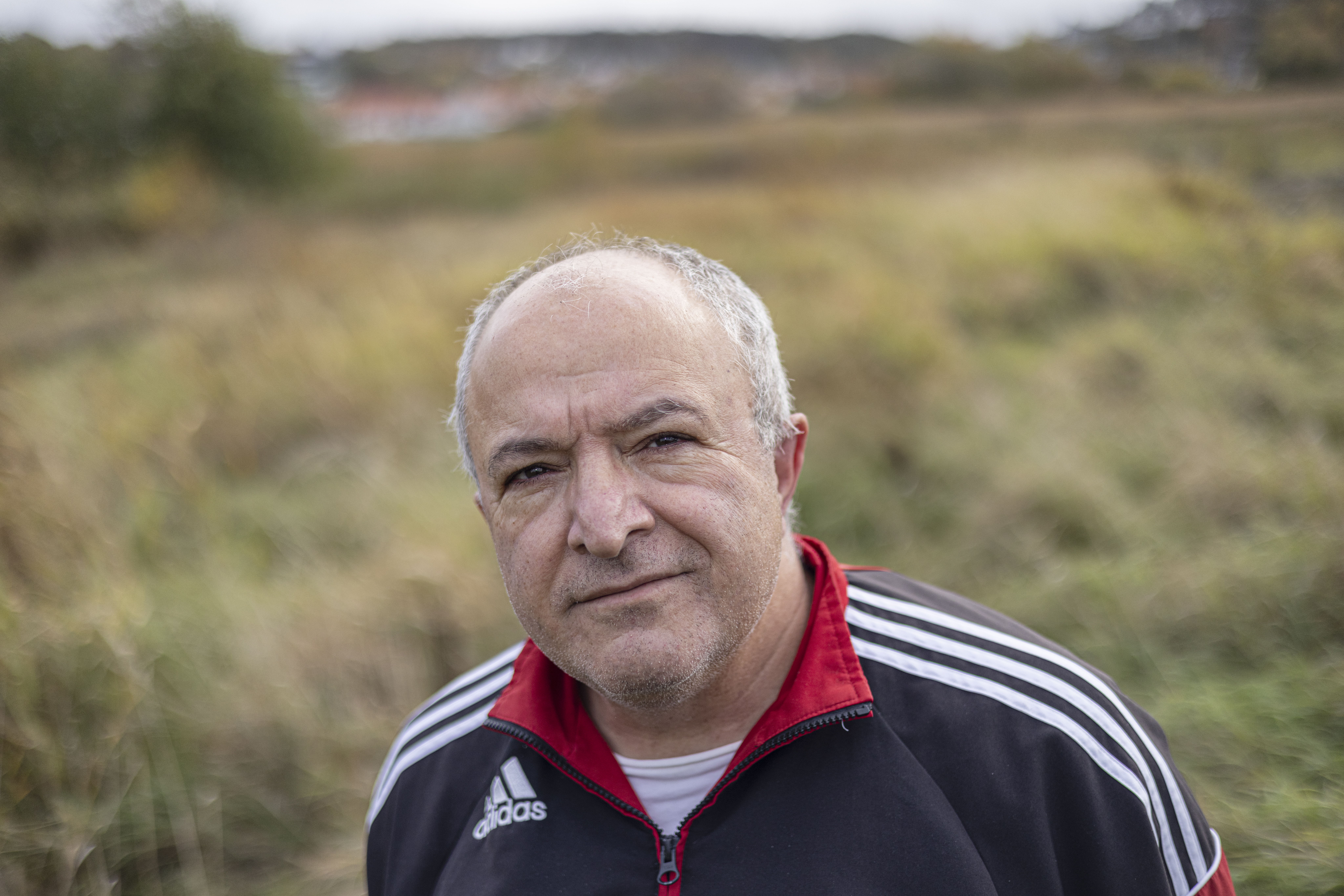 En person står utomhus iklädd en träningsjacka. I bakgrunden syns en gräsbevuxen fält och hjälpa kullar.
