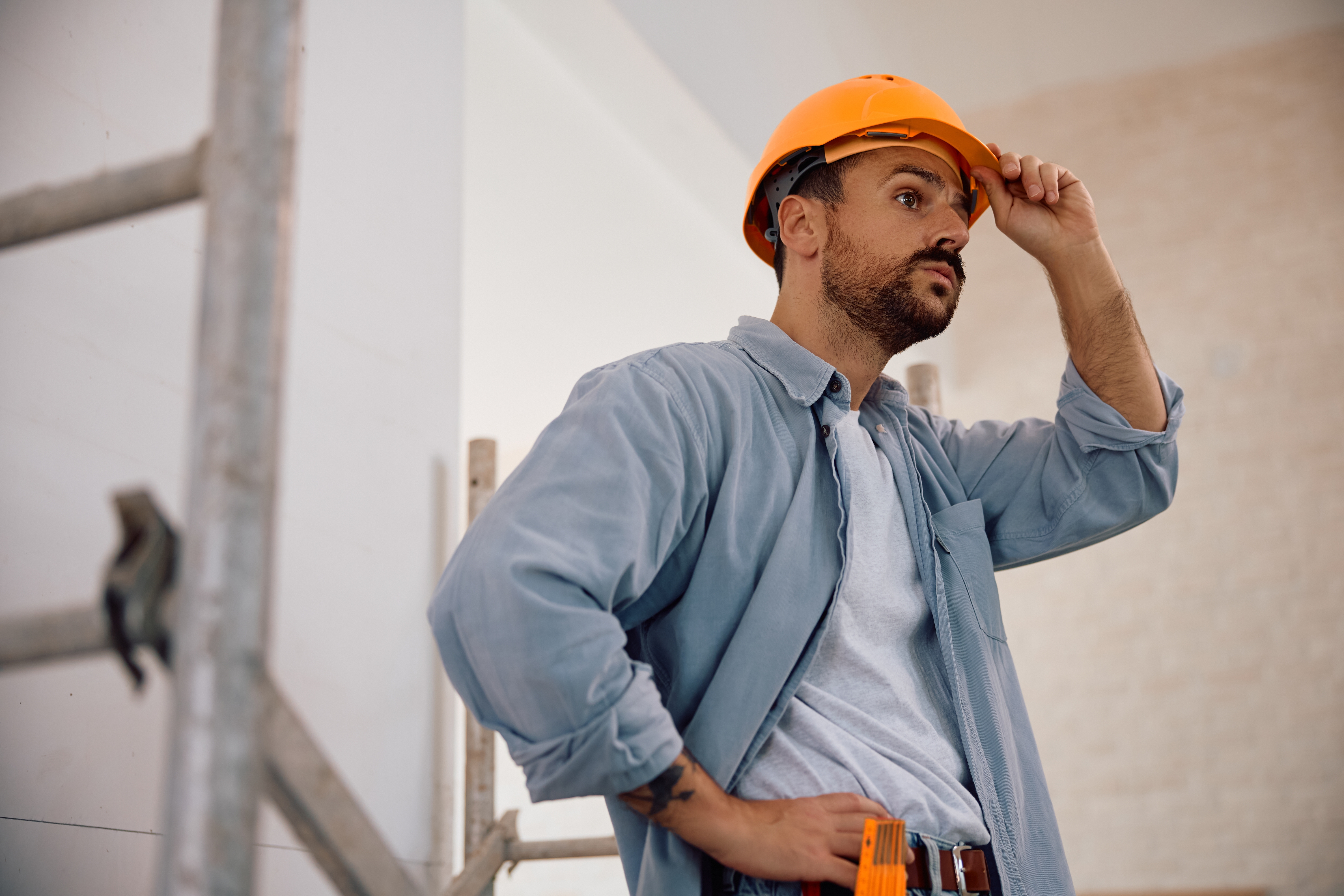 En byggarbetare i blå skjorta justerar sin orangea hjälm medan han står bredvid byggnadsställningar.