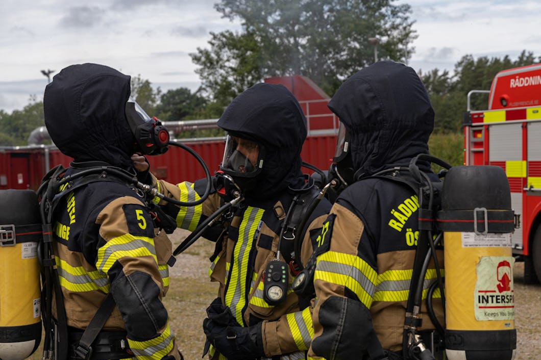 Tre brandmän i full utrustning och andningsapparat står nära en brandbil. De ser ut att förbereda sig för en insats, och en av dem justerar en annan brandmans utrustning.