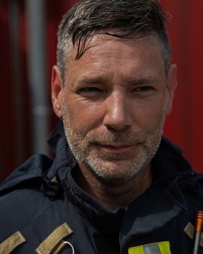Martin Bruér med kort, grånande hår och en trimmad skäggväxt, iförd en mörk brandmansuniform med reflekterande tejp, står framför en röd bakgrund.