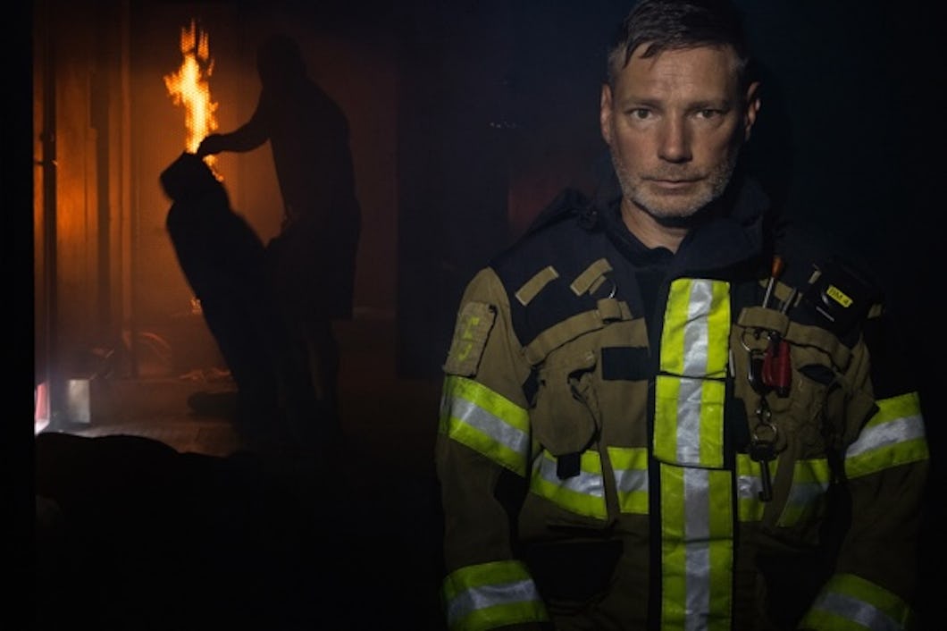 Martin Bruér i full utrustning står i förgrunden, medan en person i bakgrunden verkar rädda en annan från ett brinnande rum.