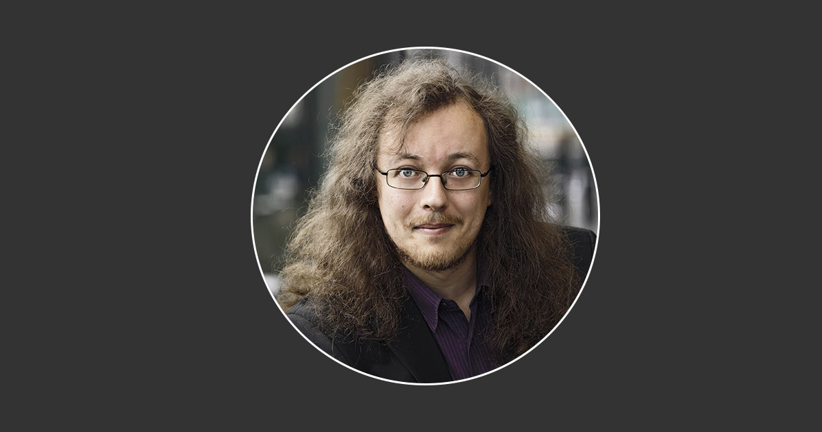 Martin Malmgren med långt lockigt hår och glasögon, iklädd en mörk blazer och en lila skjorta, stående mot en suddig bakgrund.