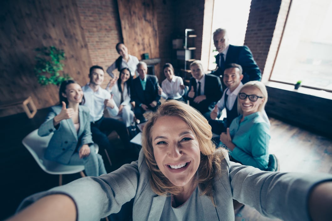 En grupp på tio personer i arbetskläder tar en gruppselfie i ett välbelyst kontor med träväggar. De ler och några gör tummen upp gester.