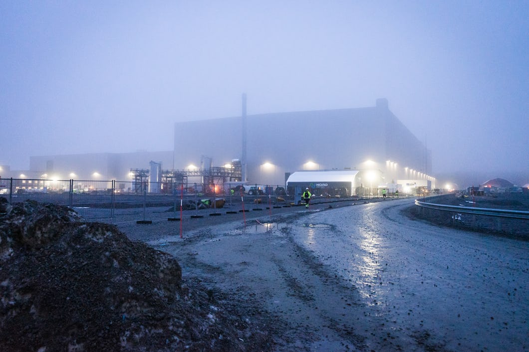 Ett stort industriellt byggnadskomplex delvis täcks av dimma. Lampor belyser strukturen och arbetare i säkerhetsutrustning syns.