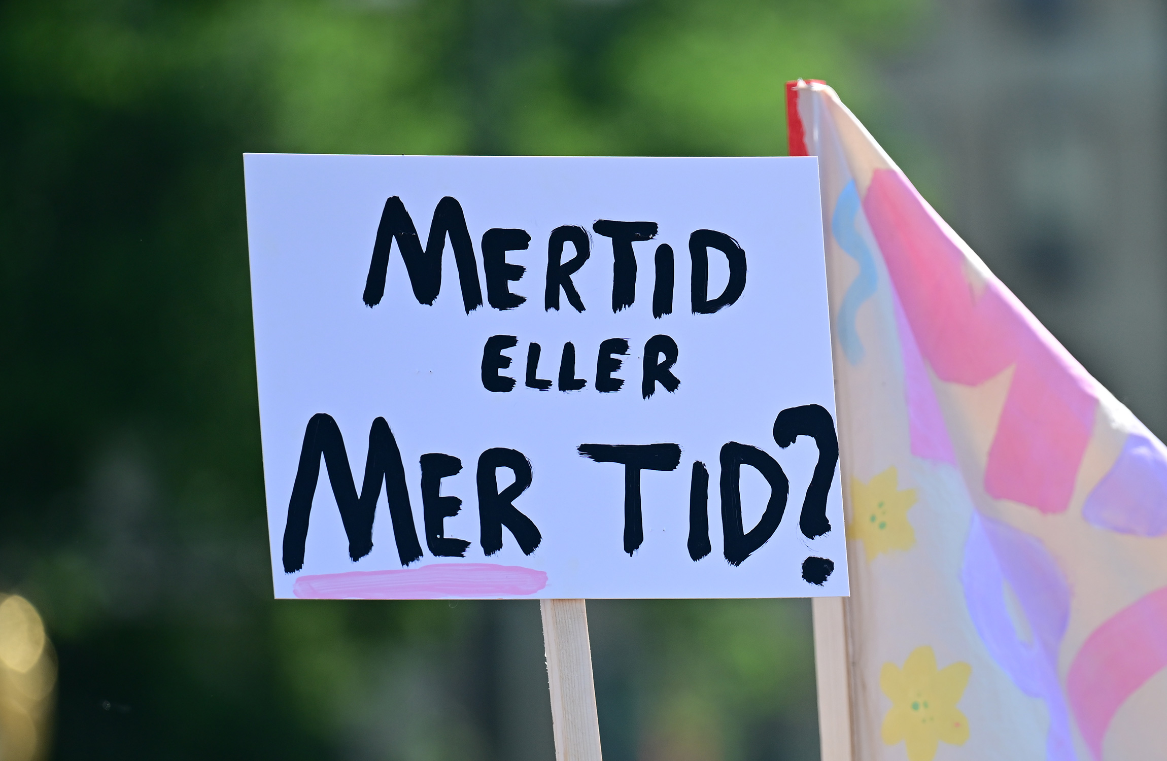 En skylt med svart text på norska lyder "MERTID ELLER MER TID?" bredvid en färgglad flagga.