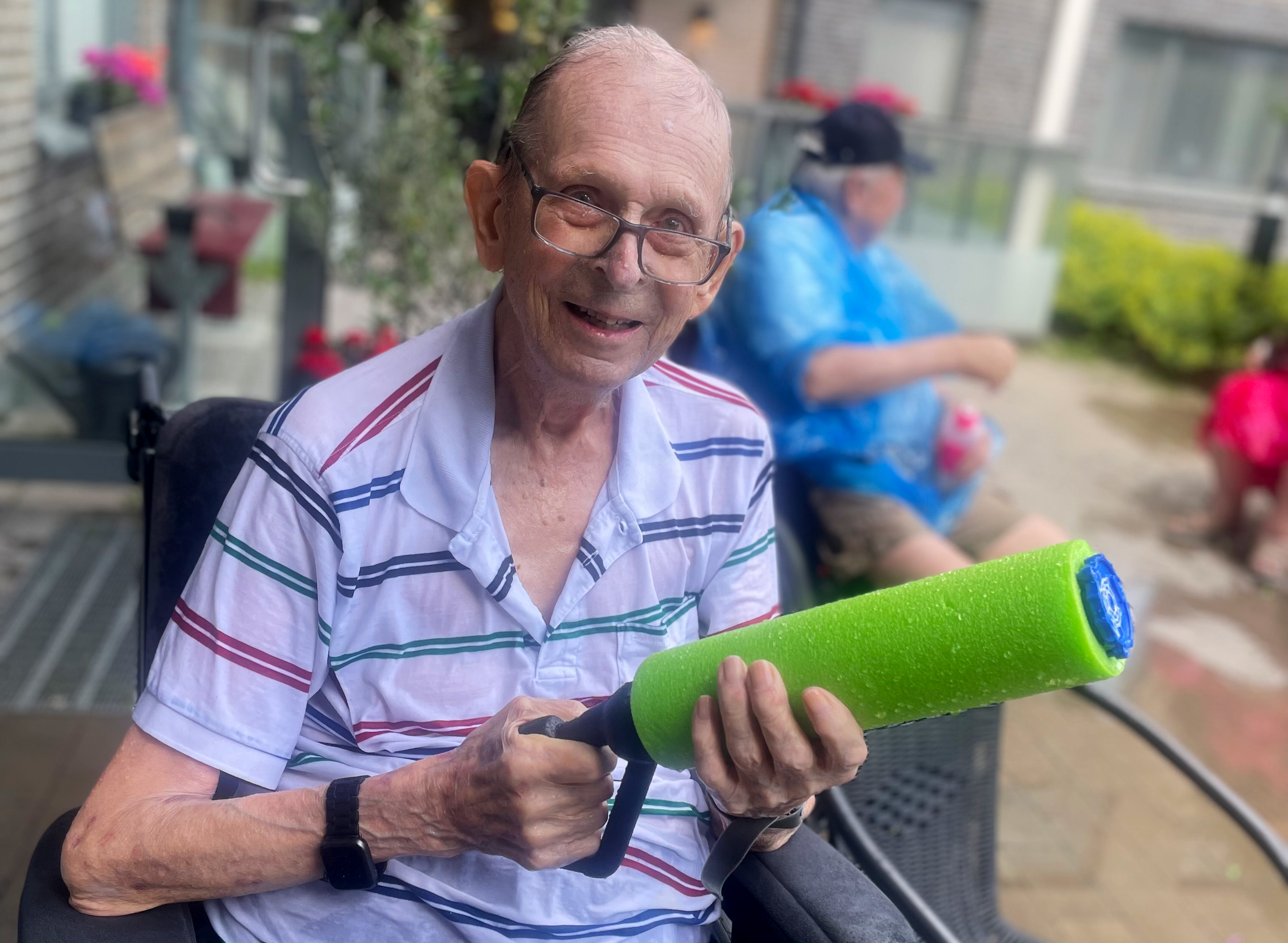 En äldre man, Gunnar Nilsson, i en randig skjorta håller i en grön skumvattenpistol utomhus. En annan person sitter i bakgrunden.
