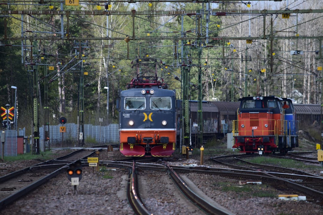 Två tåg på järnvägsspår vid en järnvägsstation omgivna av grönska och överliggande ledningar. Ett tåg rör sig framåt medan det andra står stilla till höger.