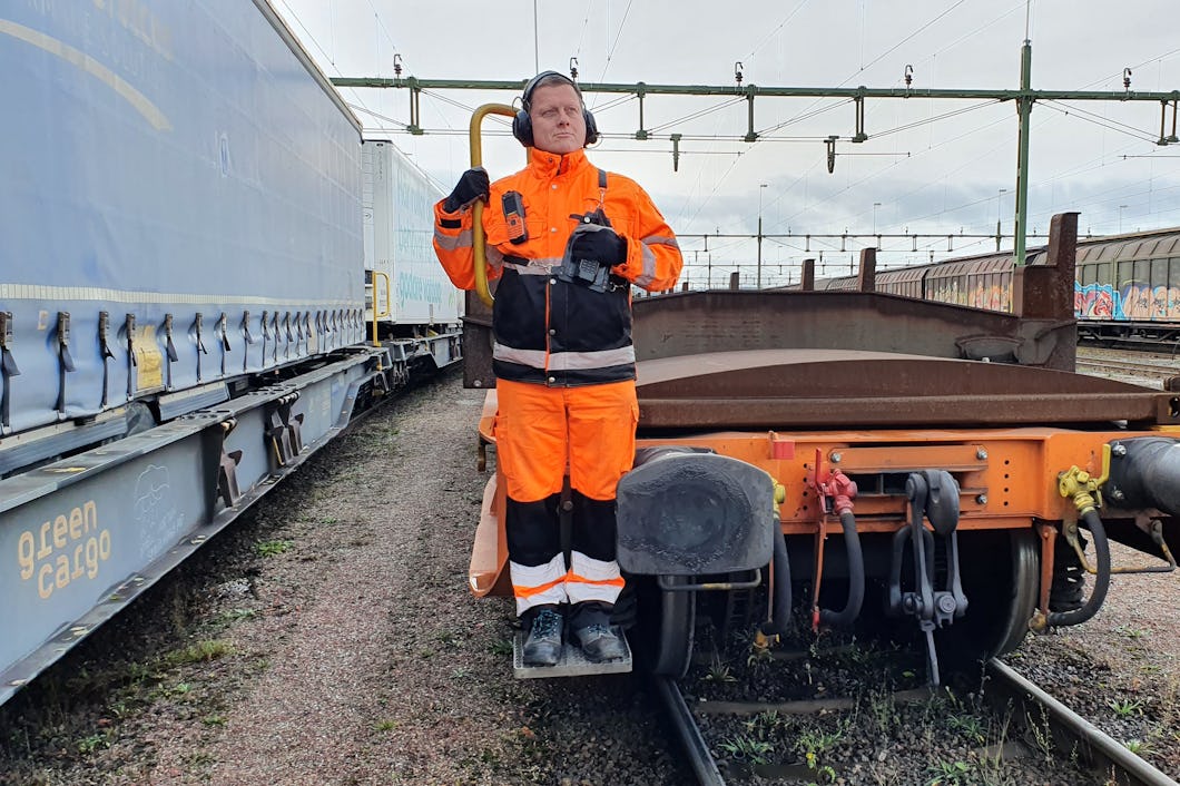 En person i orange skyddsdräkt står på ett fotsteg längst bak på en godsvagn.