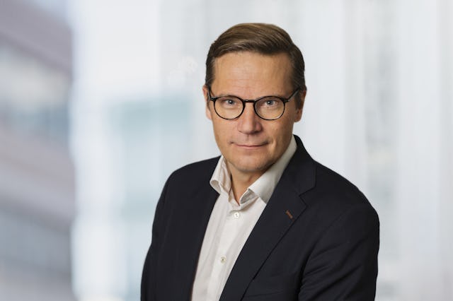 Erik Sturegård med glasögon, vit skjorta och mörk kavaj tittar rakt in i kameran. Bakgrunden består av oskarpa byggnader.
