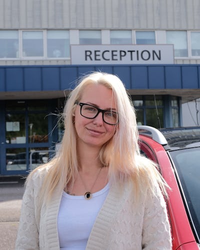 En kvinna med långt blont hår och glasögon står framför en byggnad med en skylt som säger "RECEPTION." Hon har på sig en vit topp och en ljus kofta.