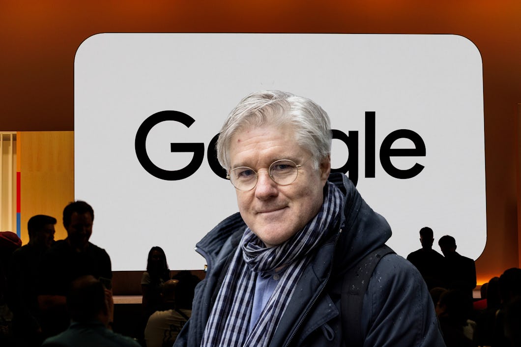 En person med grått hår och glasögon står framför en stor Google-logotyp, klädd i en mörk jacka och halsduk, vid ett evenemang eller en presentation.