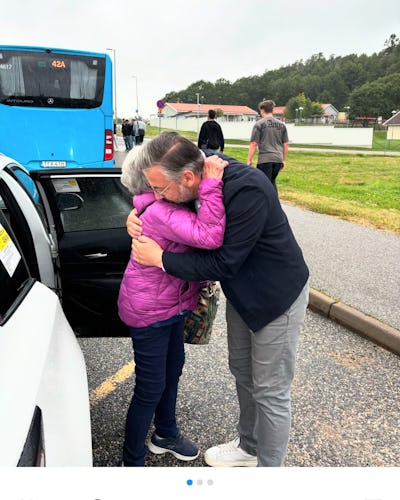 Muharrem Demiroks kramar en äldre kvinna med grått hår nära en väg, medan en blå buss och andra människor syns i bakgrunden.