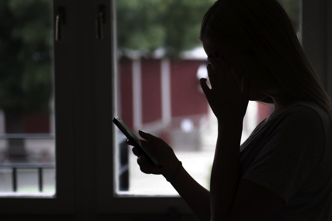 Silhuett av en person som står vid ett fönster, håller en smartphone och täcker ansiktet med ena handen.