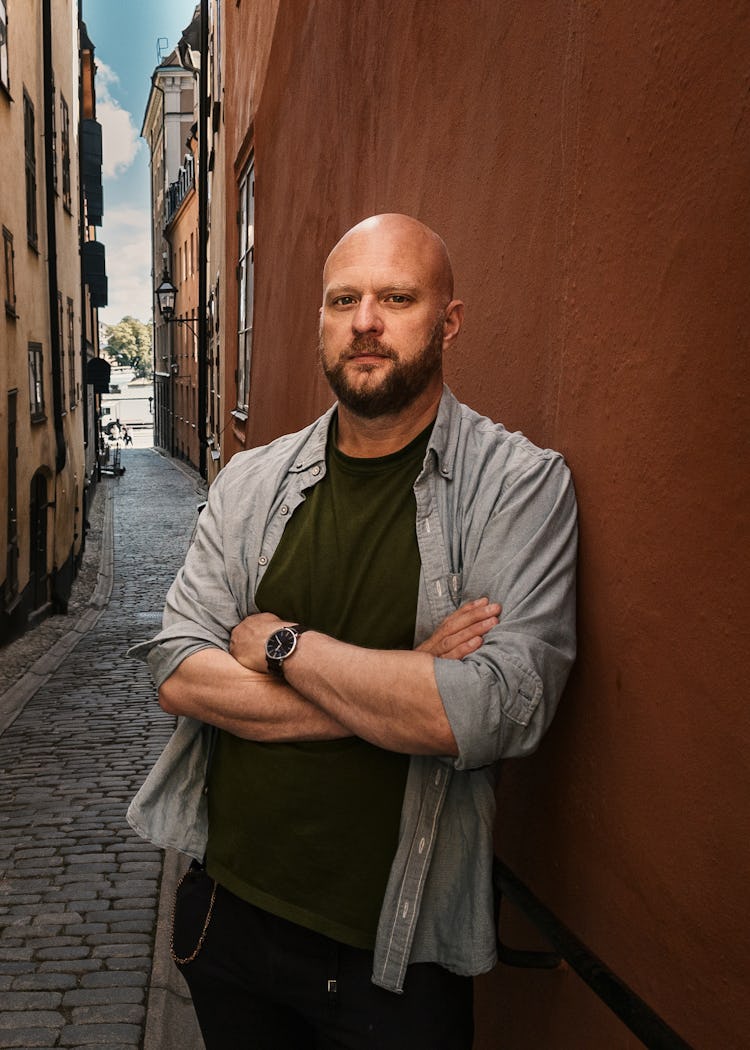 En man med skägg står med armarna i kors mot en röd vägg i en liten kullerstensgränd omgiven av gamla byggnader.