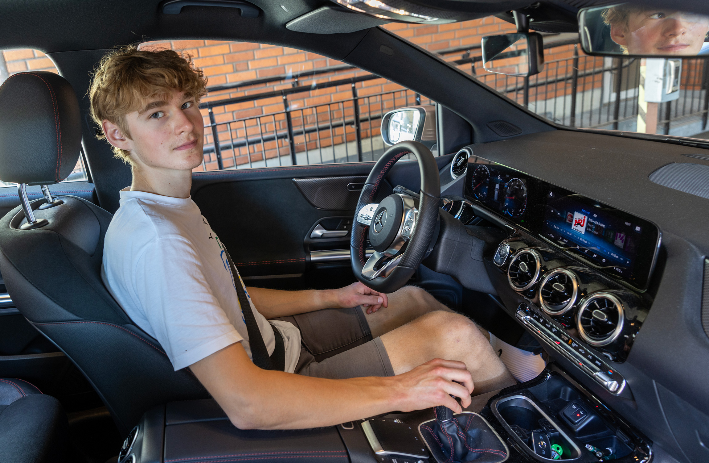 En ung person med blont hår sitter i förarsätet på en bil och tittar mot kameran.