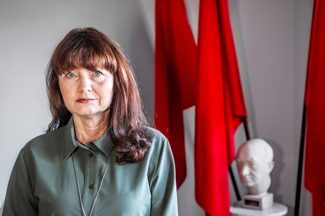 En kvinna med långt brunt hår och en grön tröja står inomhus. Bakgrunden visar röda flaggor och en vit statyett.