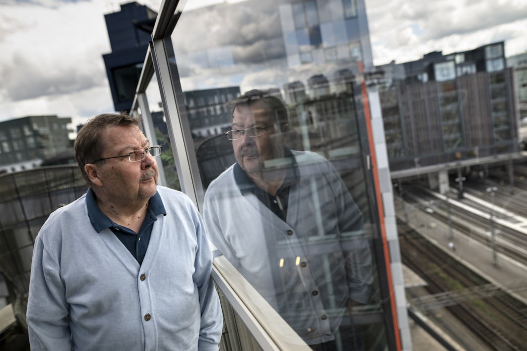Bengt Sandberg står på en balkong, bär glasögon och en ljusblå kofta, och tittar ut i fjärran. Hans spegelbild syns i glaset bredvid honom.