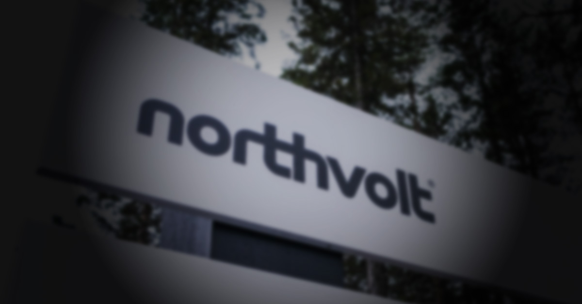 En närbild på en skylt med "northvolt" logotypen i en mörk, suddig utomhusmiljö med träd i bakgrunden.