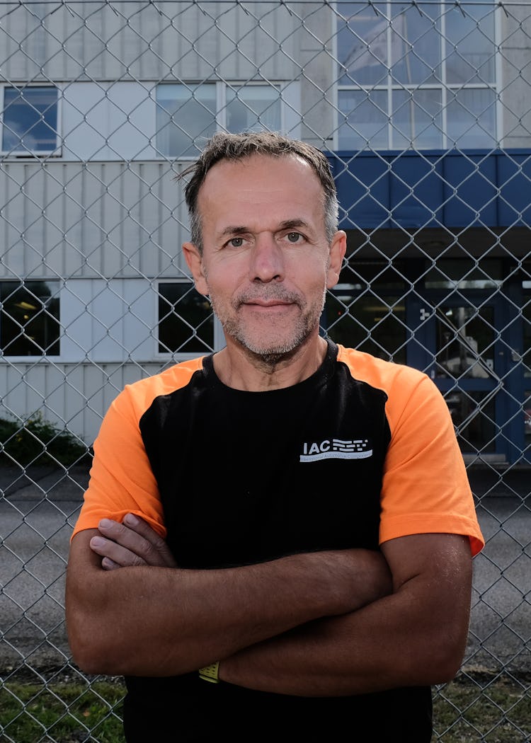 En man med kort hår och skägg, klädd i en svart och orange träningsskjorta, står med armarna i kors framför ett staket av kedjelänk och en byggnad.