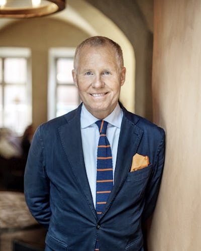 pontus sjöstrand klädd i en blå kostym, randig slips och orange bröstnäsduk står inomhus mot en beige vägg och ler.