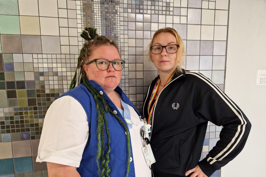 Två personer, Lina Jakobsson och Camilla Westerlund, en med grönt flätat hår och glasögon som har på sig en blå väst, och en annan med blont hår och glasögon som har på sig en svart jacka, står framför en kaklad vägg.
