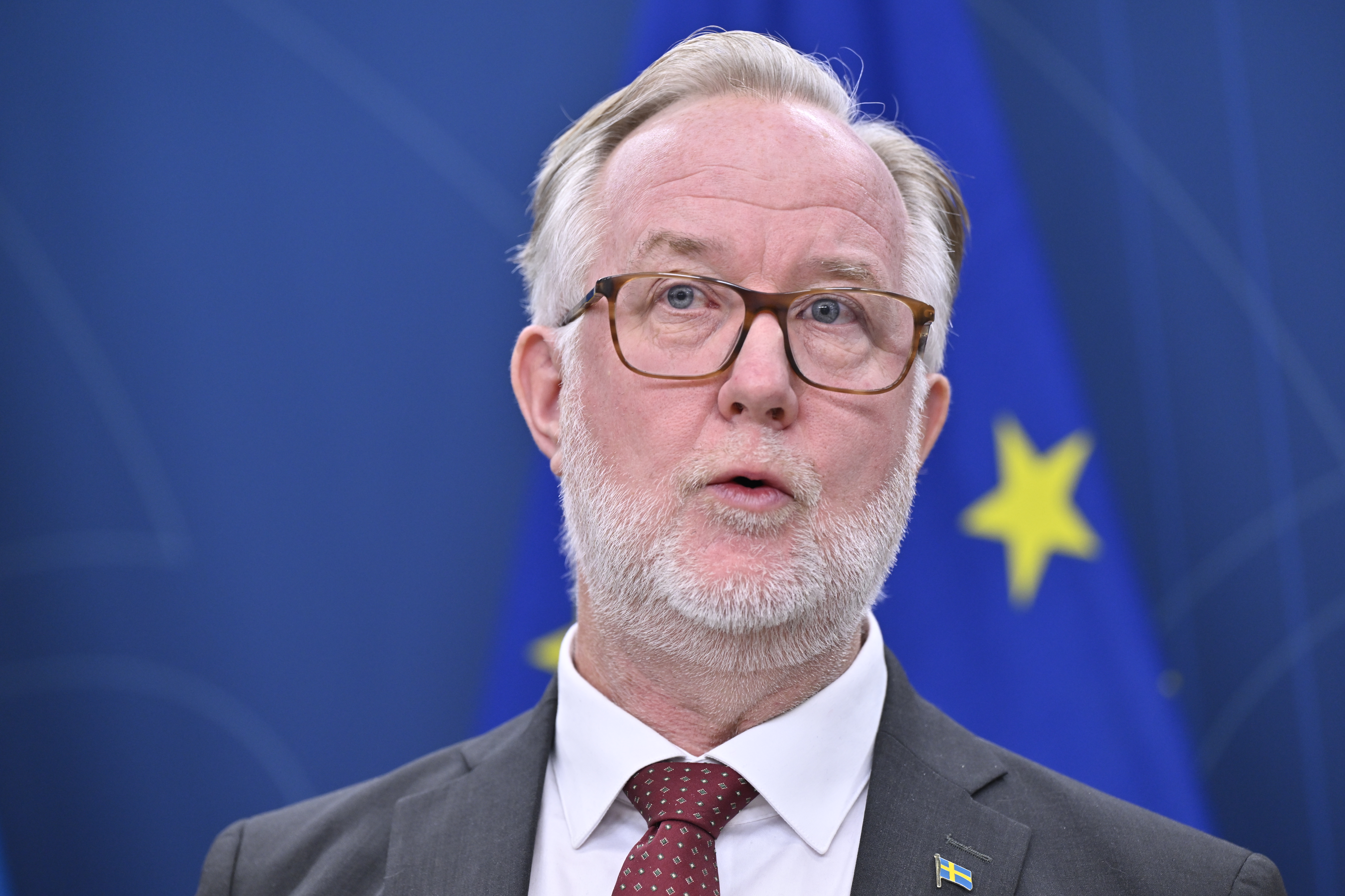 En man i kostym och slips talar framför en blå bakgrund med gula stjärnor.