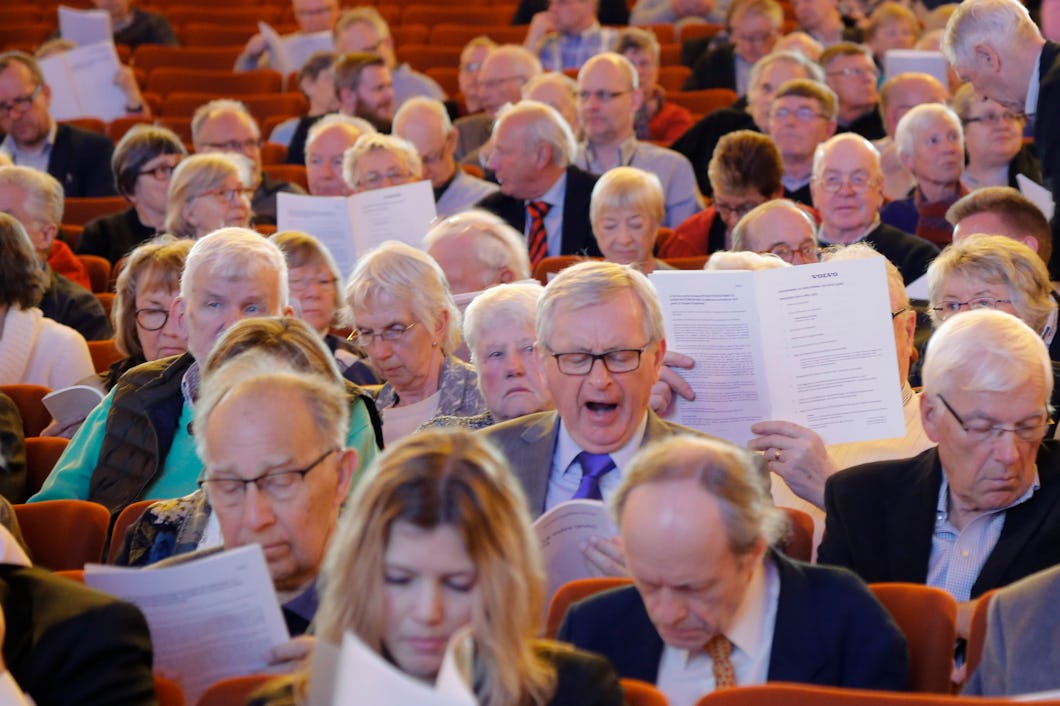 En grupp människor, mestadels medelålders och äldre, sitter i en auditorium och läser från papper och häften. Några pratar med varandra.