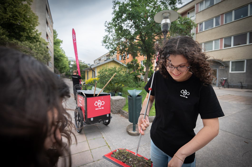 Tasnim Rabia i svart t-shirt sopar skräp in i en sopskyffel på en utomhusgång nära byggnader, med en vagn i bakgrunden och en annan person i förgrunden.
