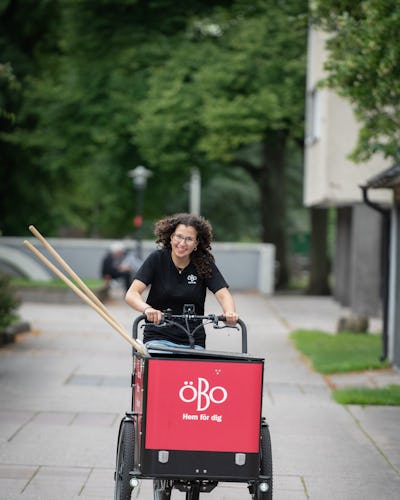 Tasmin Rabia cyklar på en röd lastcykel med texten "ÖBO Hem för dig" på framsidan, i en stadsmiljö med gröna områden.