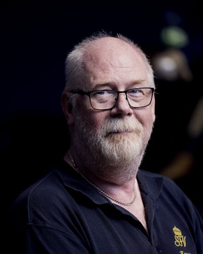 Roger Carlsson, klädd i en svart pikétröja med en broderat logotyp, står mot en mörk bakgrund.